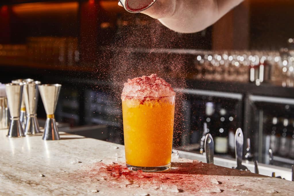 some powder being dusted over the top of a bright orange coloured drink in a glass