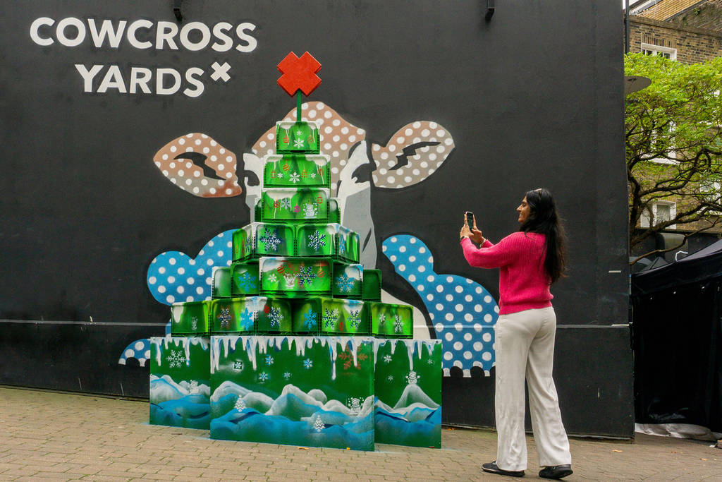 someone taking a photo of the cowcross yards christmas tree installation, made up of painted recycled crates stacked in a christmas tree shape