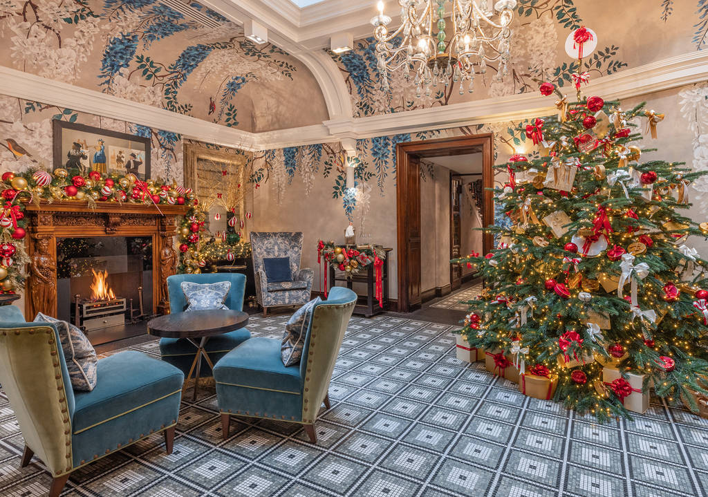 an ornate sitting room, with armchairs clustered around a side table next to a fireplace, and a charming christmas tree on the other side of the room