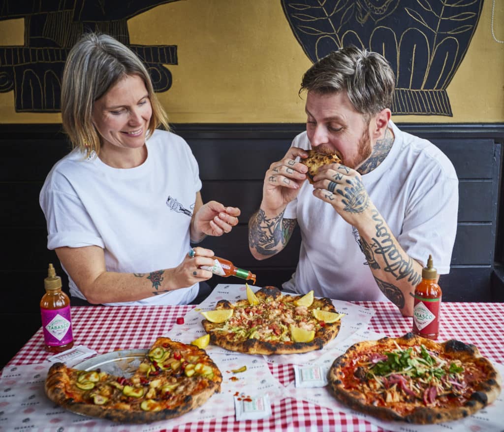 two people sitting at a table with three outrageous looking pizzas, one is about to bite into a slice, while the other is drizzling tabasco on a pizza