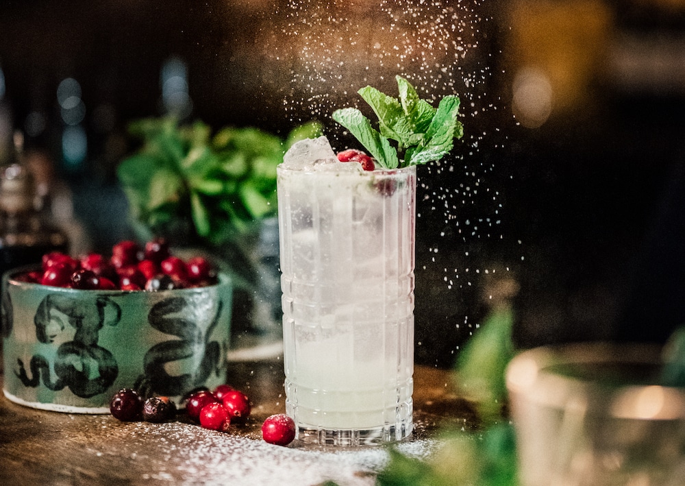a white cocktail presented in a high-ball glass with cranberry and mint garnish and snow falling over it