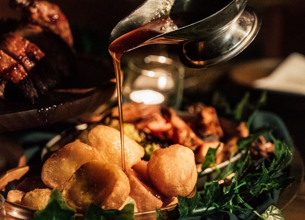 roast potatoes being drizzled with gravy, with other christmassy dishes out of focus in the background