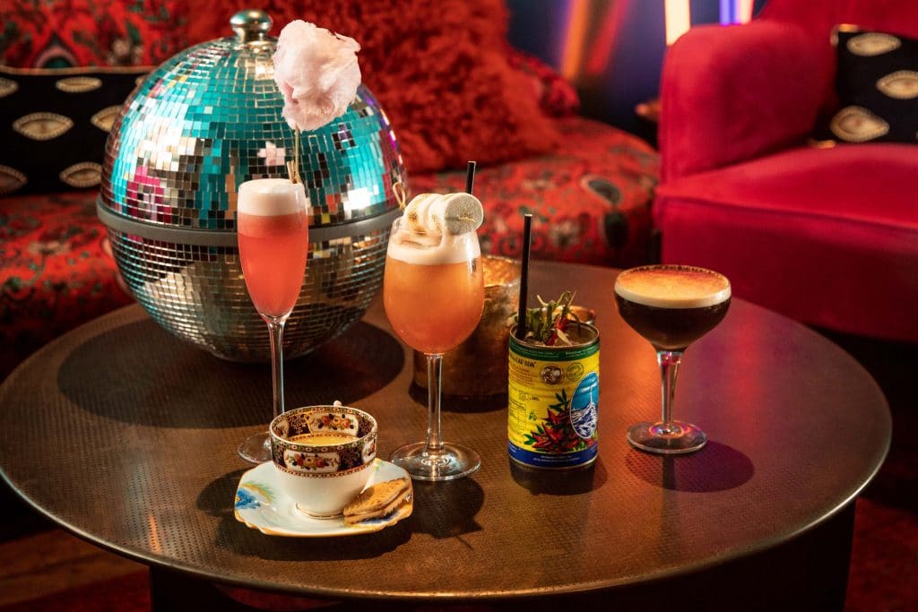 A table full of drinks in front of a disco ball and a red sofa, at The Little Scarlet Door.
