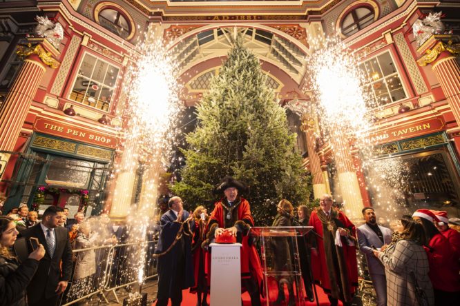 the lord mayor turning on the lights at leadenhall market, flanked by fireworks going off, in front of the christmas tree
