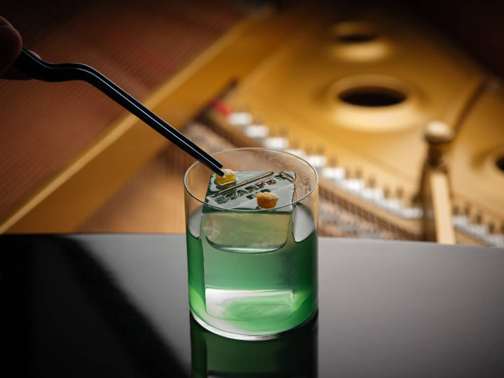 a chartreuse green cocktail with a scarfe's embalzoned ice cube, with someone tweezing a small garnish onto the ice cube