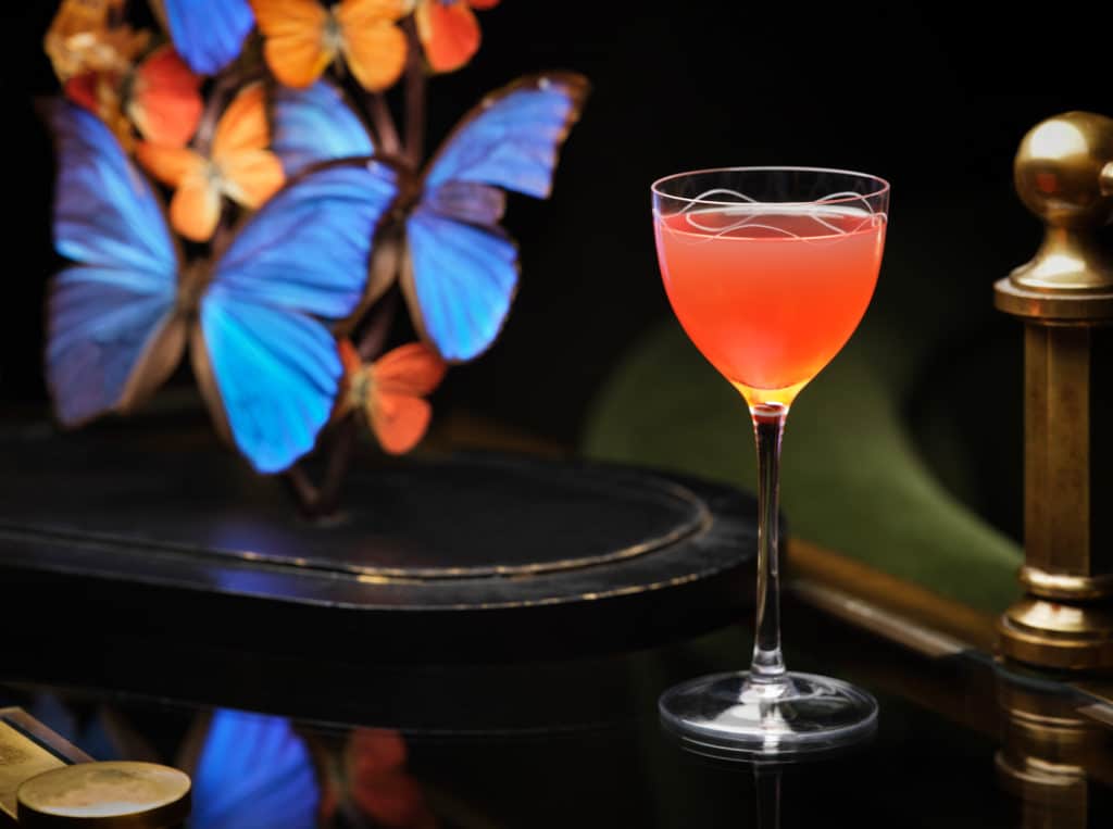a grapefruit pink hued cocktail in a nick and nora glass, next to a display of butterflies
