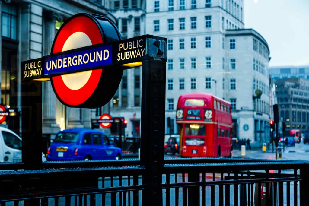 An entrance to the London Underground, with a red, London bus driving by.