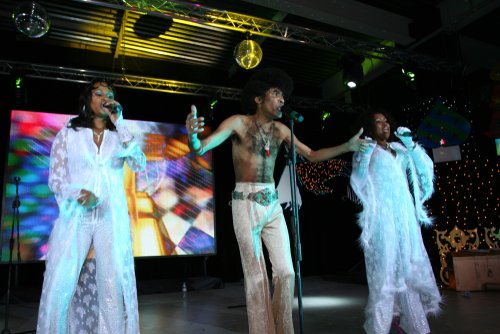 Three of the singers off Boney M performing at a gig to a crowd of people surrounded by bright lights 