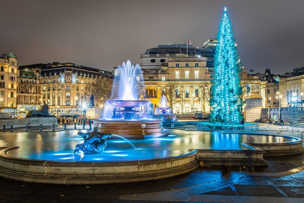 the trafalgar fountain at night, lit with a blue hue, with a magnificent christmas tree in the background