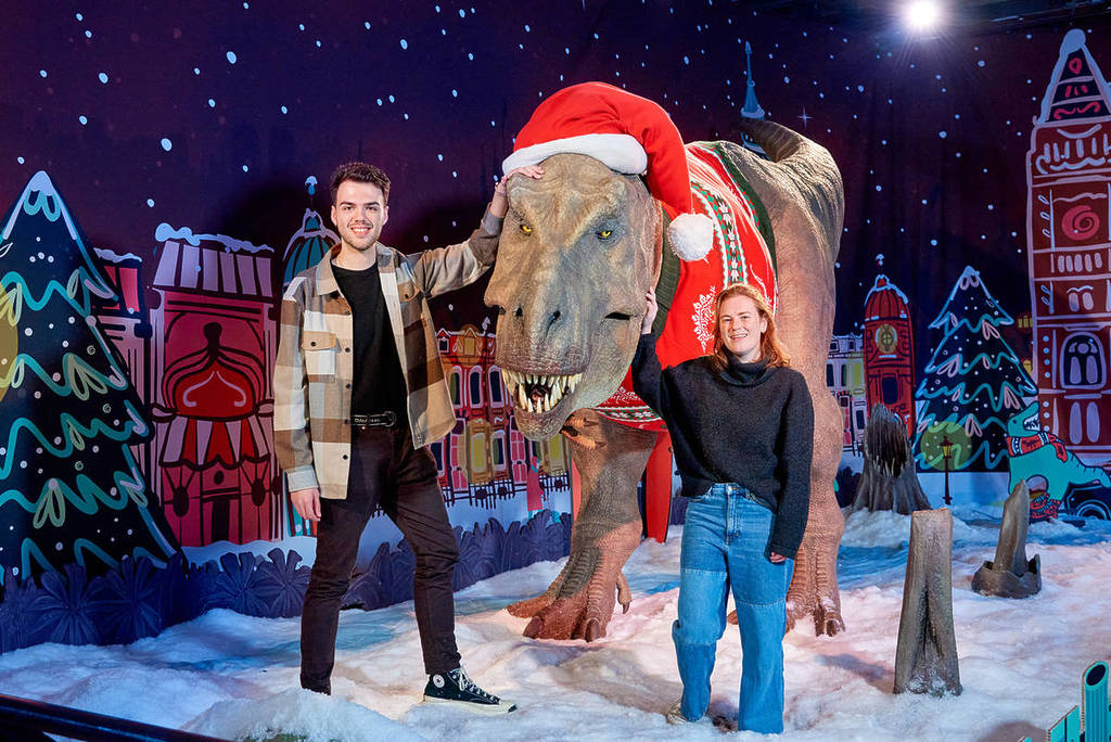 two people posing alongside the natural history museum's t-rex, which has been kitted out in a christmas sweater and hat