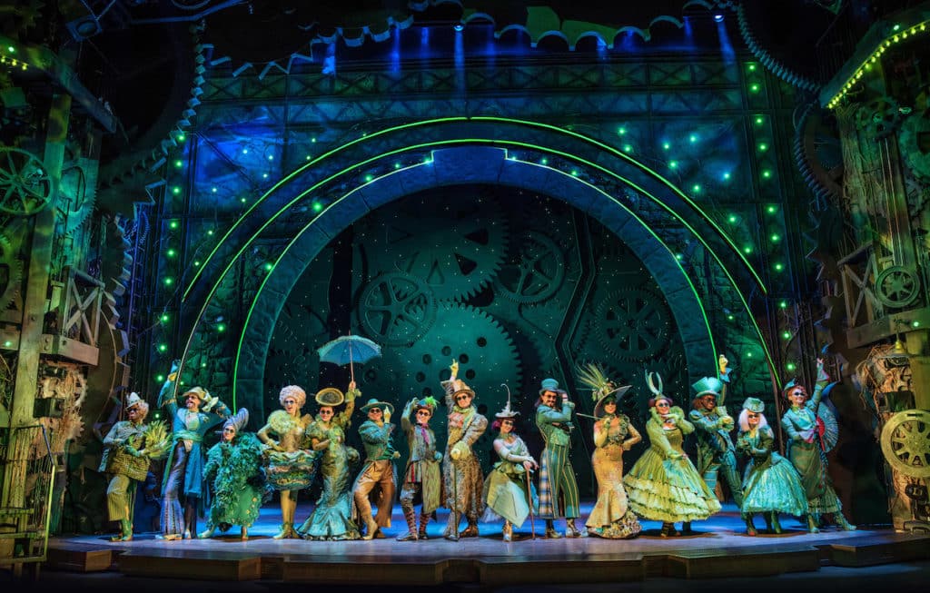 the cast of Wicked, all dressed in glamorous green costumes, line up of stage with theatrical poses
