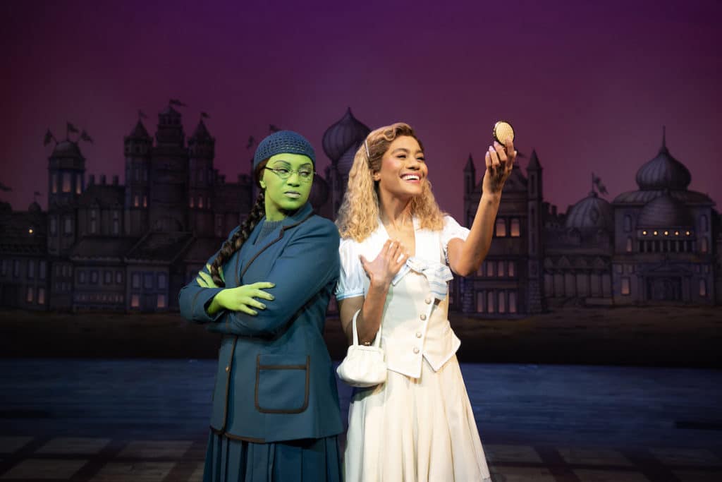 the green-skinned frowning Elphaba and blonde smiling Glinda stand back to back on stage while Glinda looks into a compact mirror