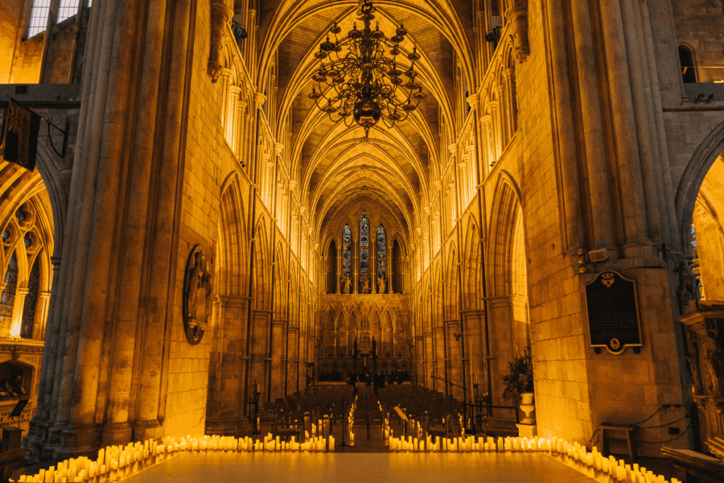 The interior of Southwark Cathedral in London bathed in the glow of candles with a chandelier hanging from the ceiling and candles lining a stage in the foreground.