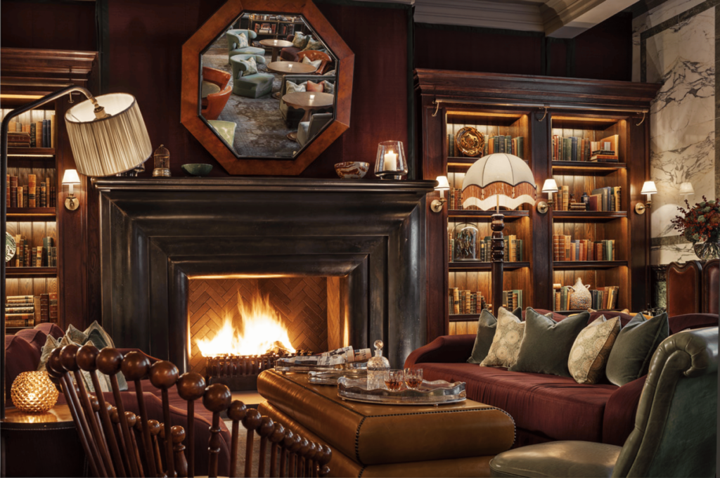the interior of the scarfes library bar, showing a roaring fire in between bookshelves, with gorgeously opulent armchairs and sofas in front