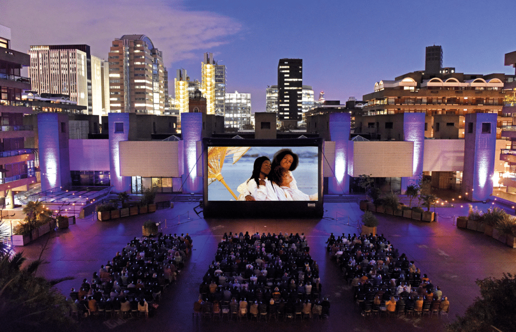 an evening outdoor screening of a film, with soft purple lighting suffusing the area as guests watch a movie on a massive screen. Various London buildings are visible behind