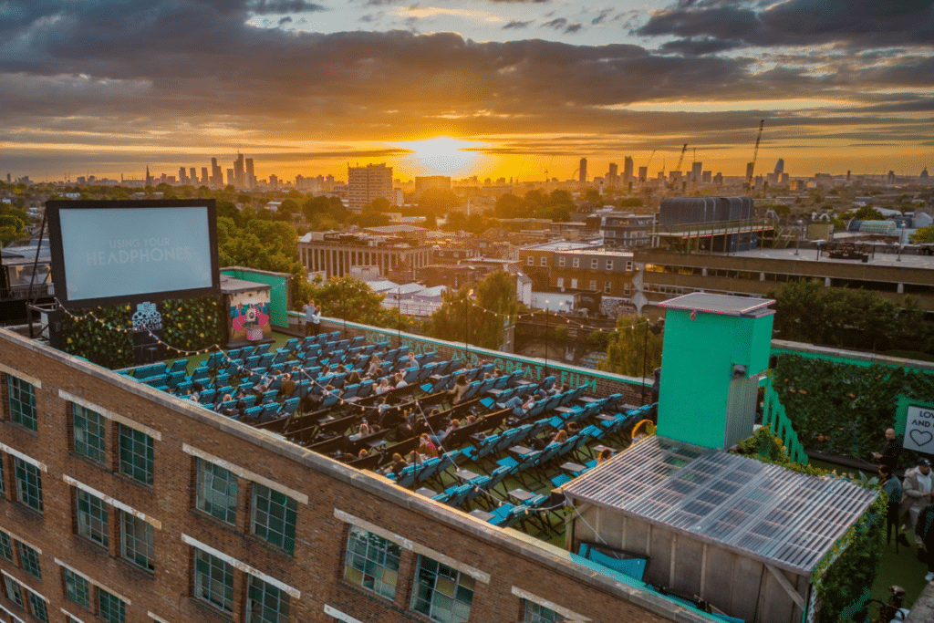 A panoramic view of the rooftop film club above the bussey building, with views over london behind it and the sun setting
