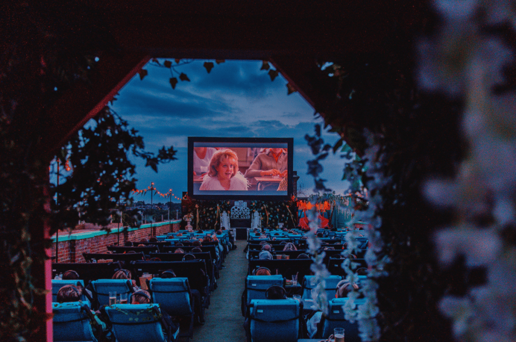 a dusk shot of the rooftop film club from behind the seats, with a film playing on the screen featuring drew barrymore